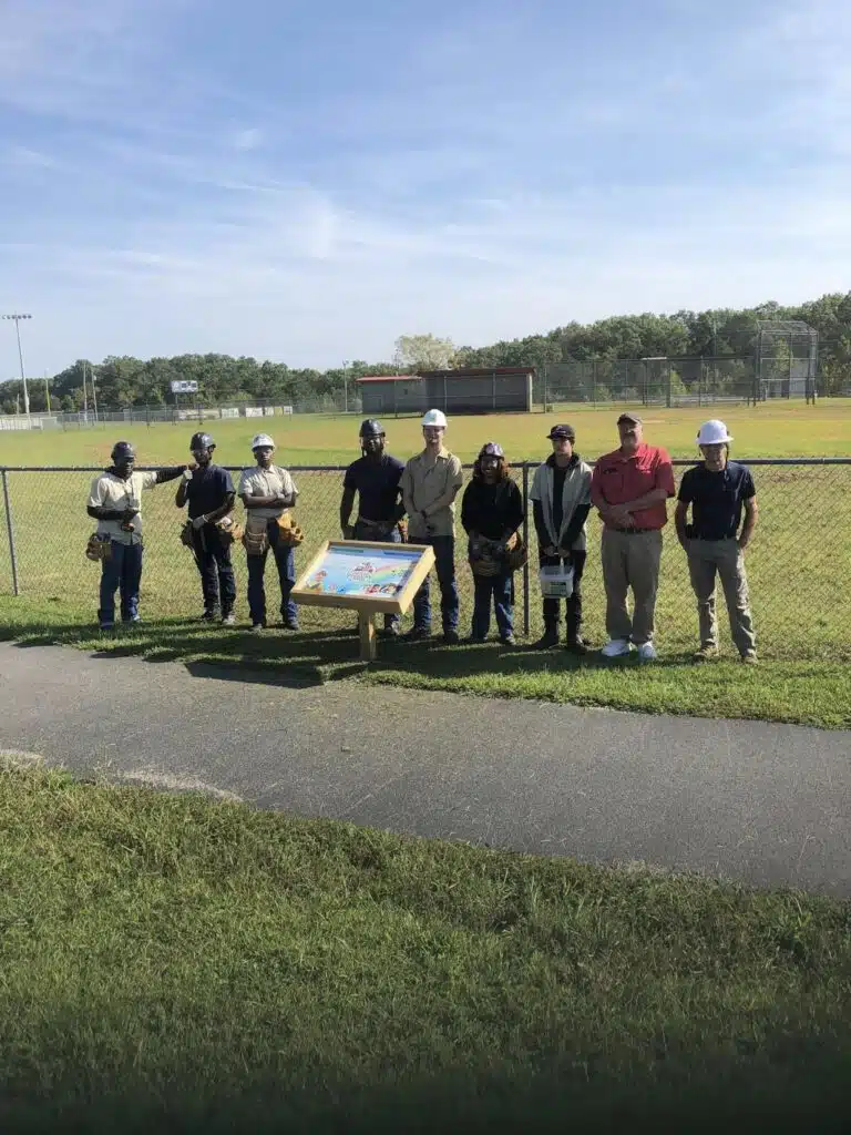 Background is green grass and sky at the local park. 9 Construction people standing by a book stand.